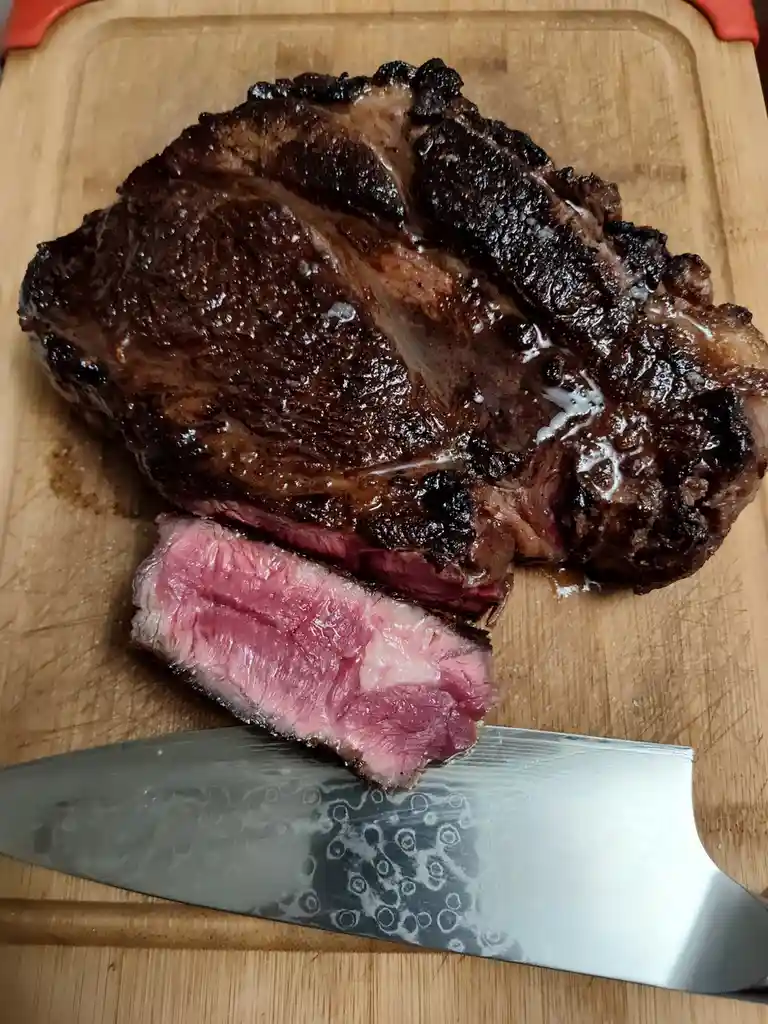 A close-up photo shows a large, seared steak on a wooden cutting board with a slice cut off, revealing a pink, medium-rare…