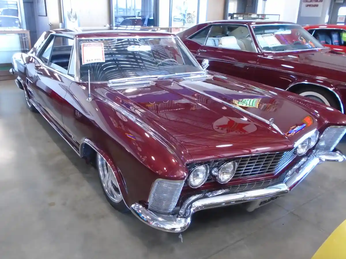 A shiny maroon classic car, likely a Buick, is displayed in a showroom with a price tag of $64,995 visible on its…