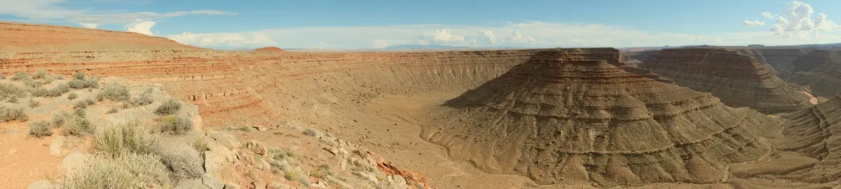 A wide-angle desert canyon panorama with layered red rock formations, sparse shrubs, and a blue sky with scattered clouds…