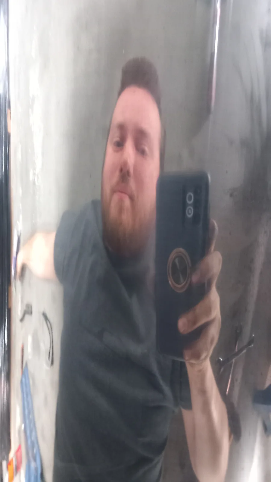 A man with a beard takes a blurry mirror selfie holding a smartphone with a circular mount, looking directly at the camera…