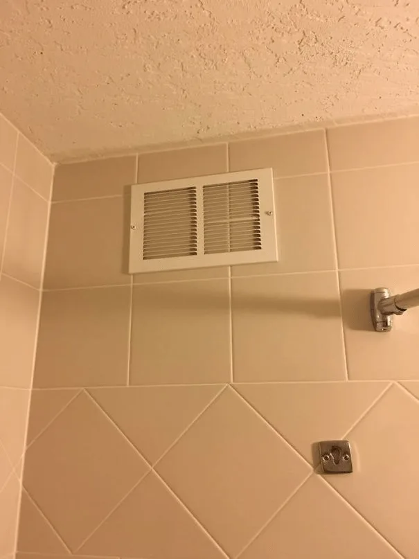 A close-up photo of a white rectangular vent mounted on beige tiled bathroom walls, with a shower arm and electrical outlet…