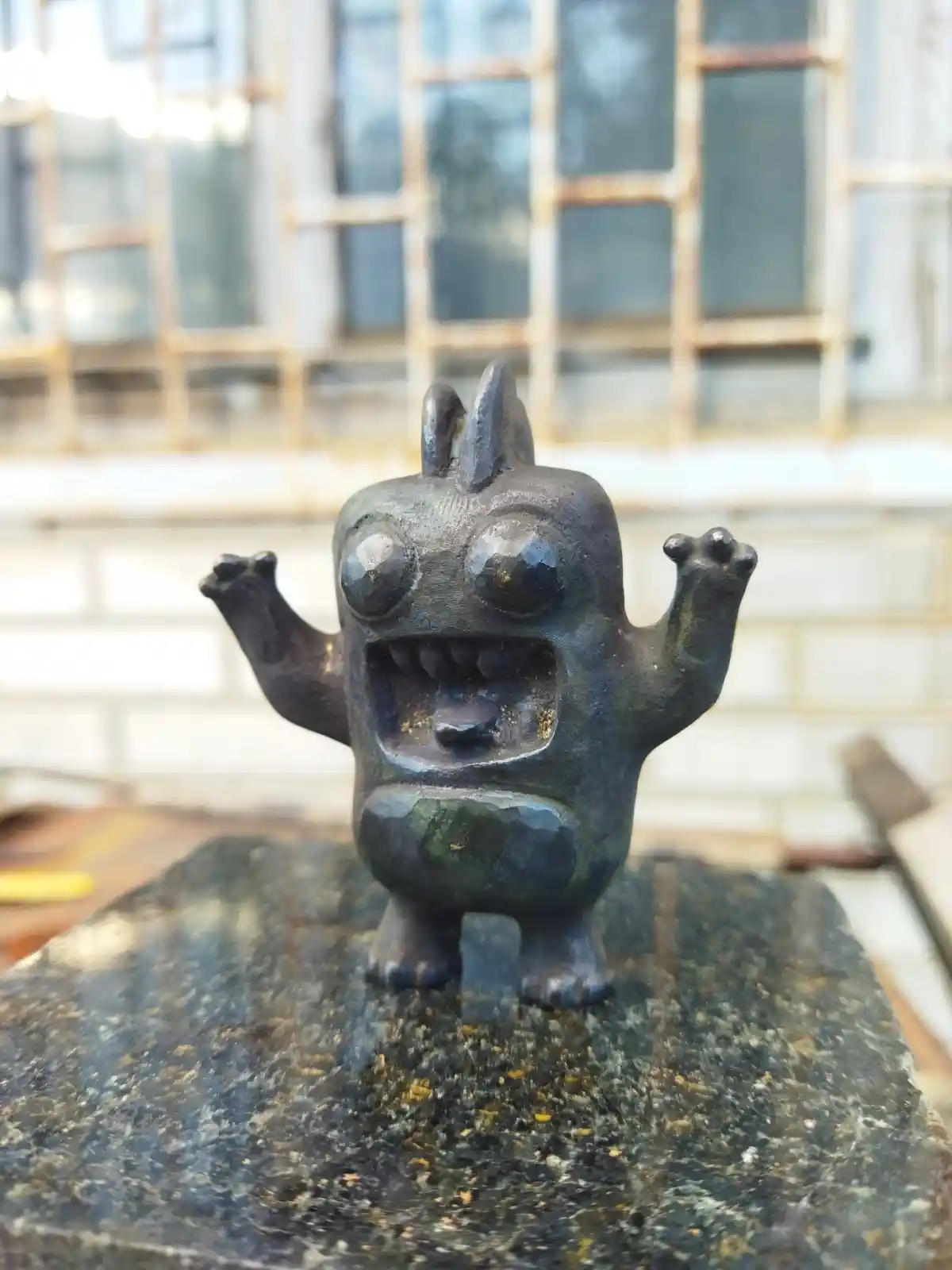 A small, dark bronze statue of a cartoonish monster with wide eyes, an open mouth showing teeth, and raised arms stands on a…