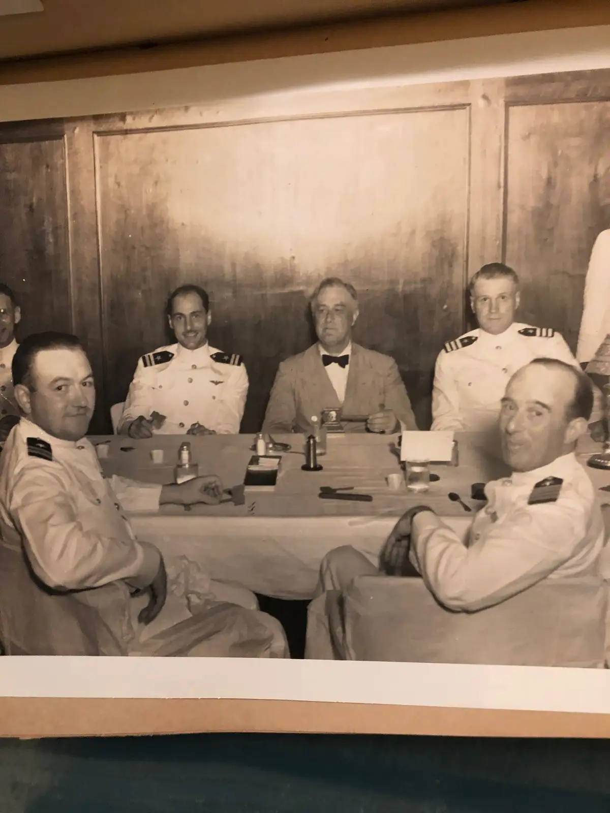 A black-and-white photograph shows a group of men in military uniforms and one man in a suit with a bow tie seated around a…