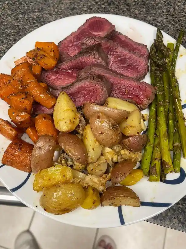 A white plate with blue trim holds a generous serving of sliced medium-rare steak, roasted carrots, roasted baby potatoes…