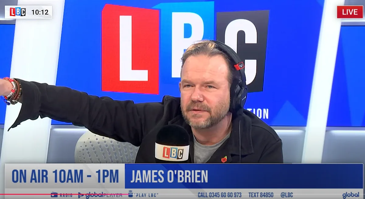 A man with a beard and headphones, wearing a black jacket over a grey shirt, sits in a studio with a large 'LBC' logo behind…