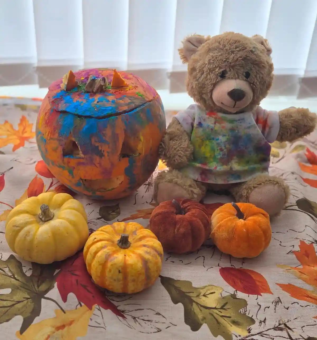 A brown teddy bear wearing a paint-splattered apron sits beside a large, colorful, paint-covered jack-o'-lantern with carved…