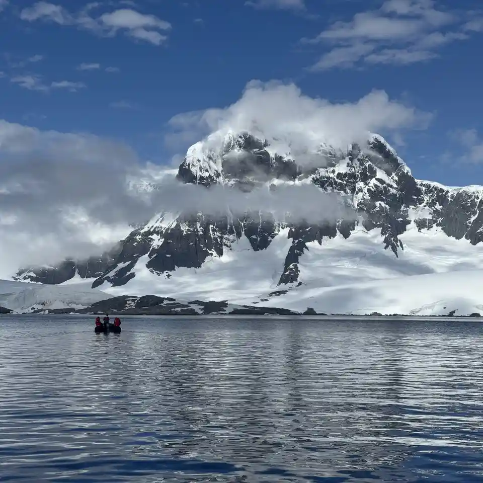 A small inflatable boat with two people in red jackets floats on calm blue water, facing a massive, snow-capped mountain…