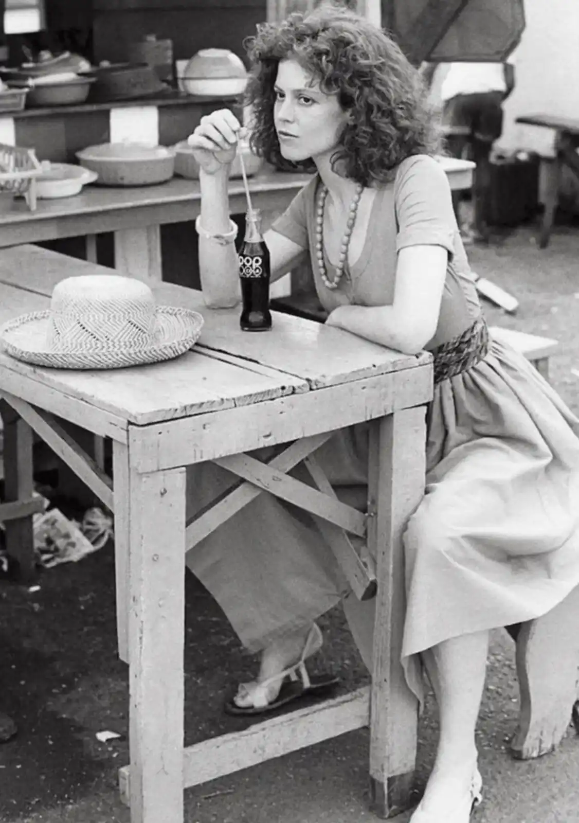 A black-and-white photo shows a woman with curly hair sitting at a wooden table, holding a straw in a soda bottle. A straw…