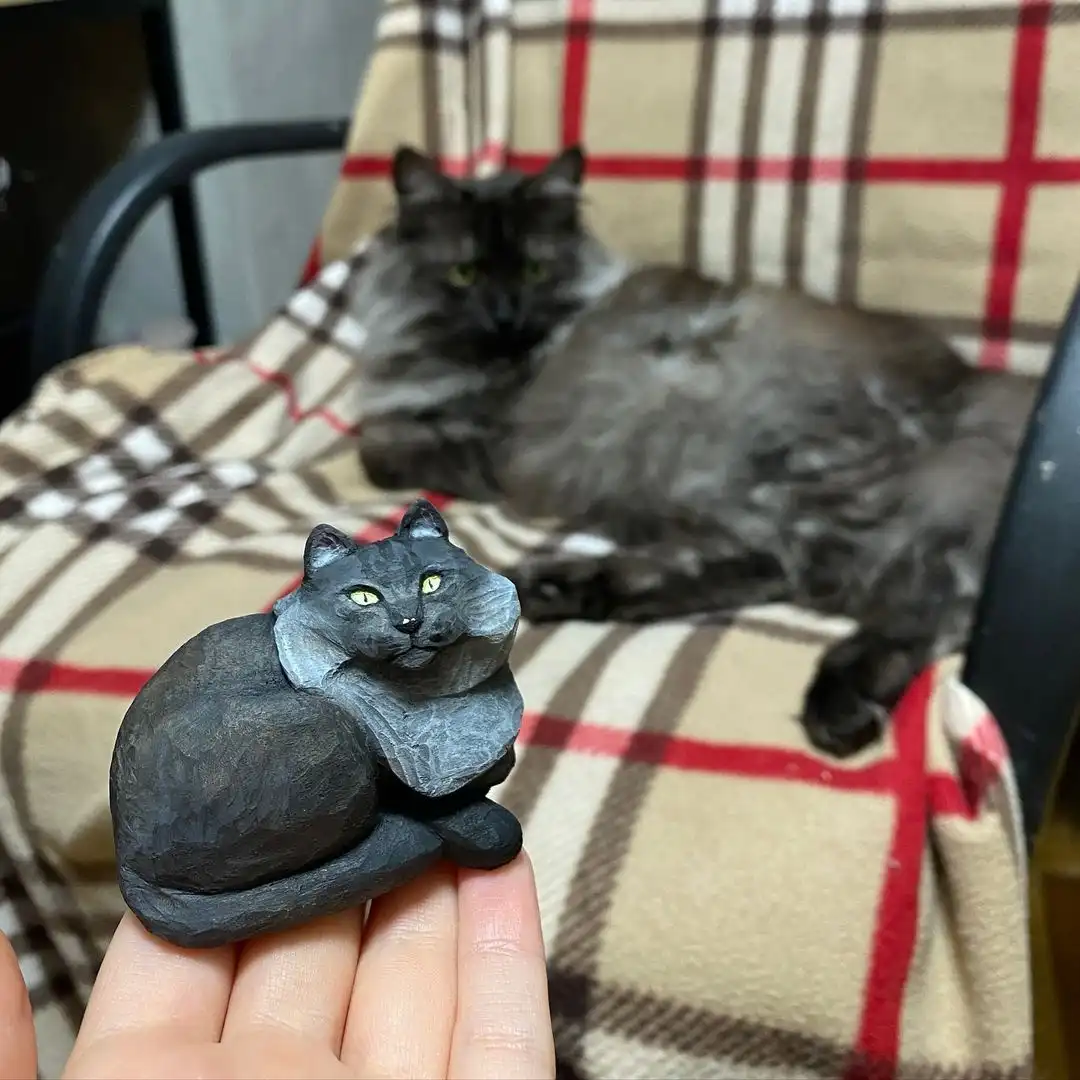 A hand holds a small, dark gray carved cat figurine with bright yellow eyes in the foreground. Behind it, slightly out of…