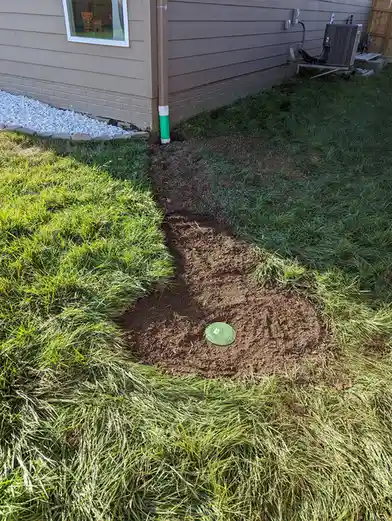 A grassy yard shows a freshly dug trench leading to a green sprinkler system cover, with a house wall and white gravel…