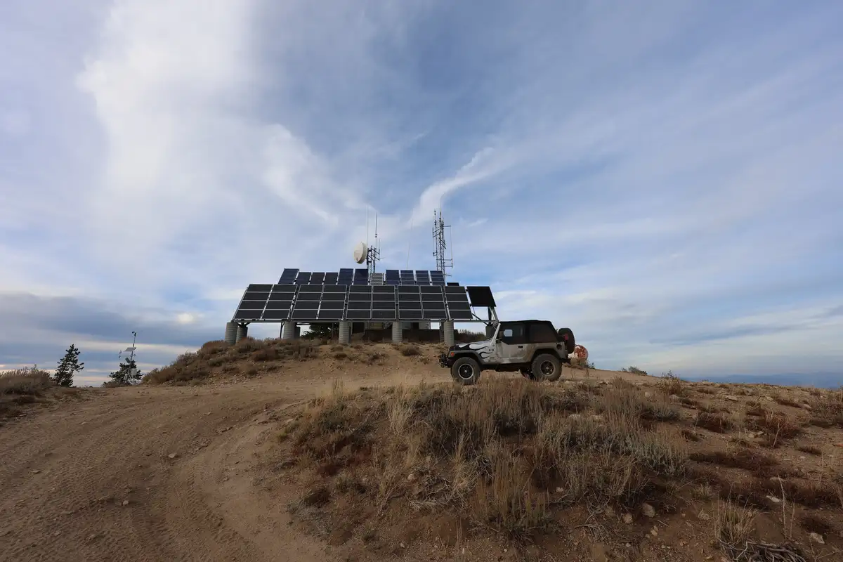 A rugged, off-road Jeep Wrangler is parked on a dirt hillside next to a large array of solar panels and communication…