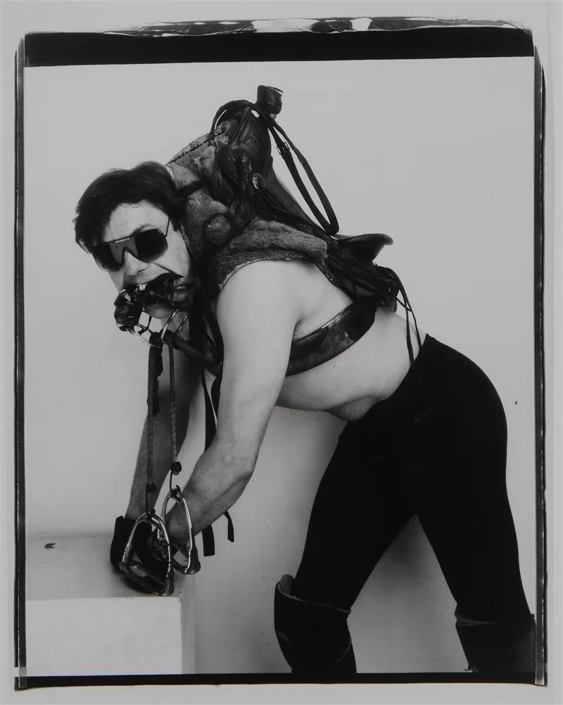 A black-and-white photo shows a man in a horse saddle, wearing sunglasses and a harness, with his mouth gagged and hands…