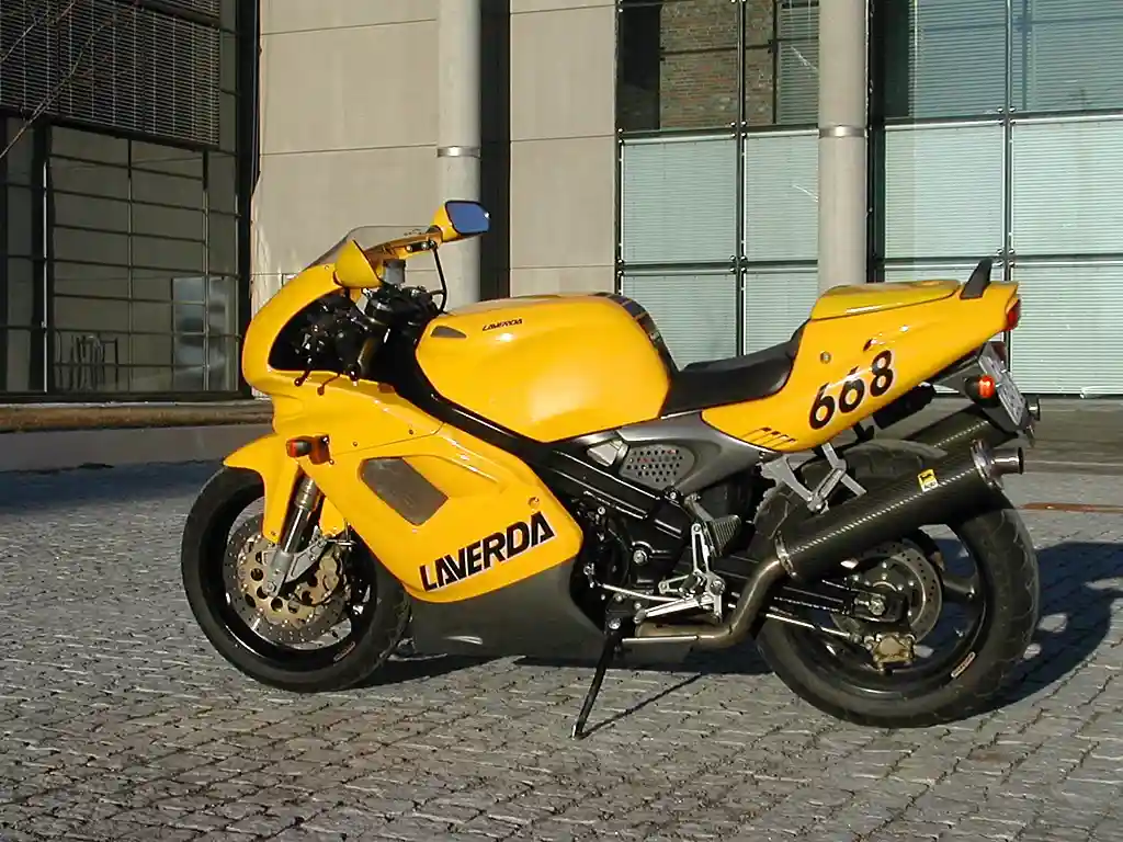 A bright yellow Laverda sport motorcycle with the number 668 on its side is parked on a cobblestone street in front of a…