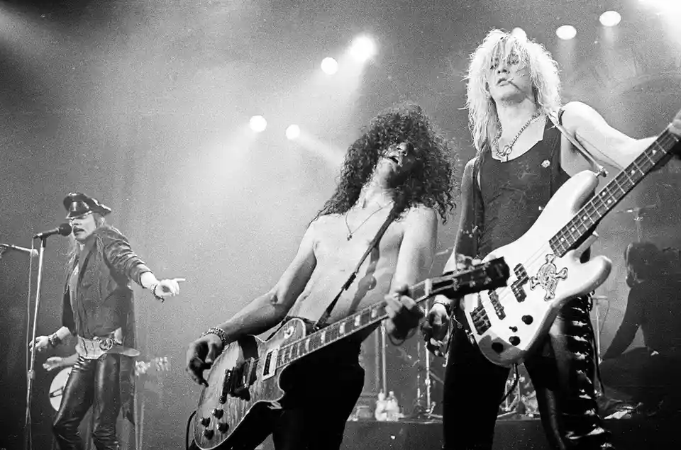 A black-and-white photo of the band Guns N' Roses performing live on stage, featuring lead singer Axl Rose on the left…