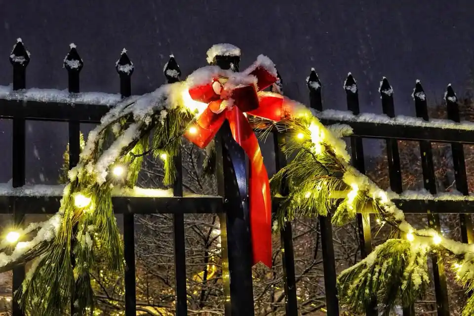 A festive Christmas wreath with a large red bow and glowing string lights is draped over a black metal fence, covered in…
