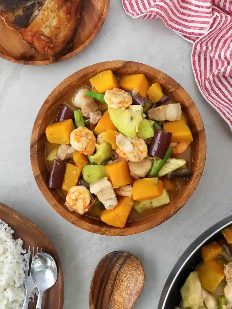 An overhead shot of a wooden bowl filled with a colorful stir-fry containing shrimp, cubed orange squash, green beans, and…