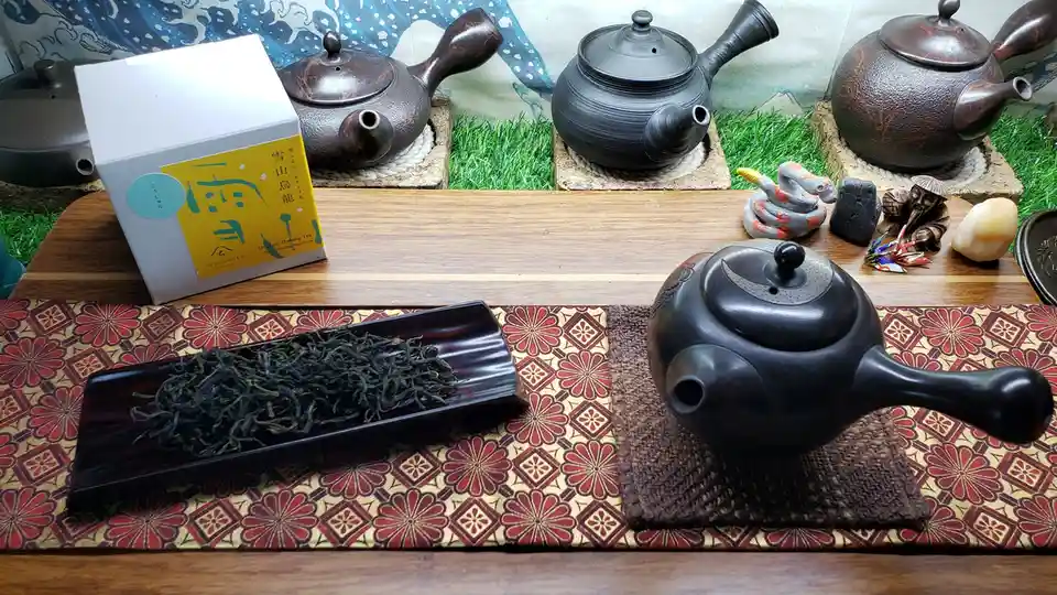 A still life of a traditional tea setup with multiple dark ceramic teapots on wooden trays, a box of 'Organic Oolong Tea'…