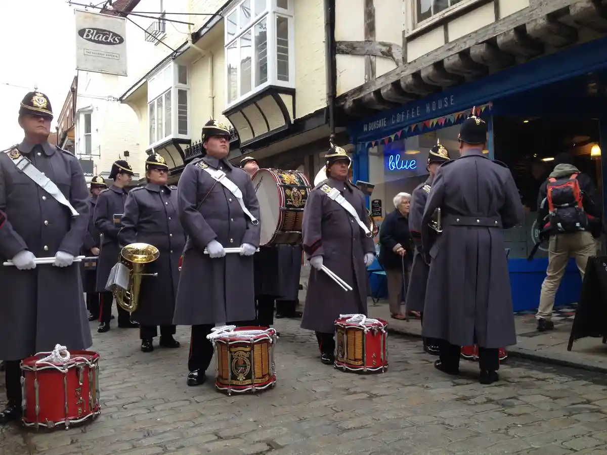 A marching band in formal grey uniforms stands on a cobblestone street in front of a blue storefront with a neon 'blue'…