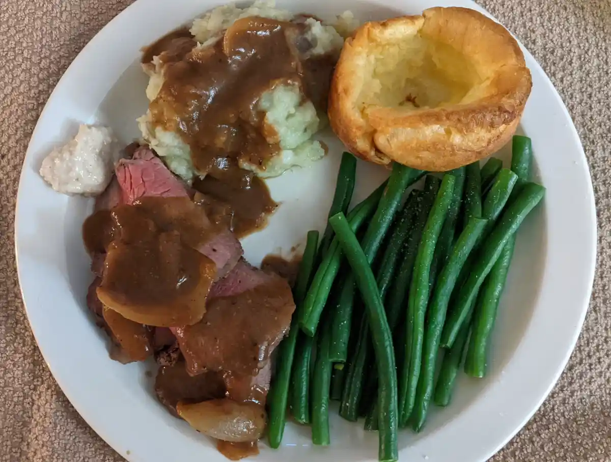 A plate of traditional roast dinner featuring sliced roast beef with gravy, mashed potatoes with gravy, green beans, a…