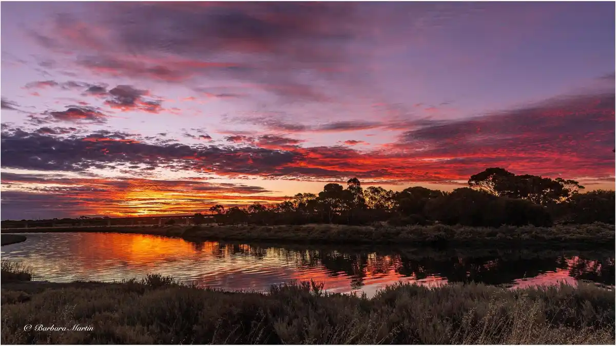 A serene landscape photograph captures a vibrant sunset over a calm body of water, with fiery red, orange, and purple clouds…