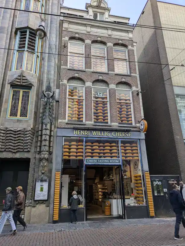 A street-level photo of the Henri Willig Cheese shop in Amsterdam, where the entire facade is covered in rows of large…