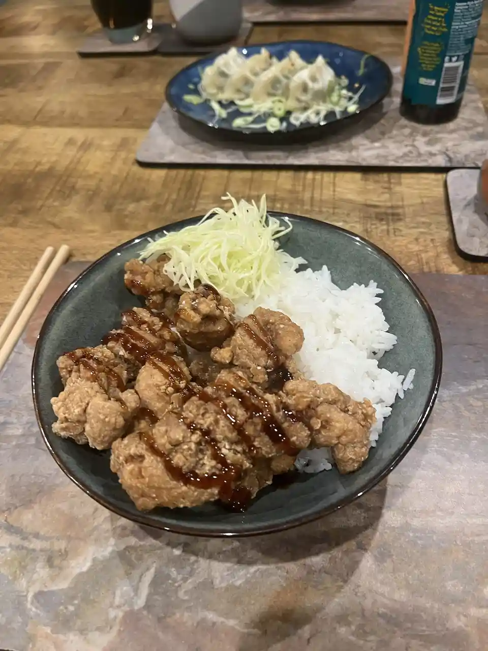 A close-up photo shows a dark green bowl filled with fried chicken pieces drizzled in sauce, served over white rice with…