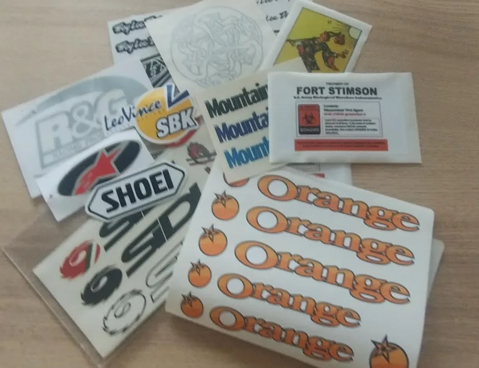 A pile of various stickers on a wooden surface, including motorcycle brands like Shoei and Leovince, a 'Mountain' sticker…