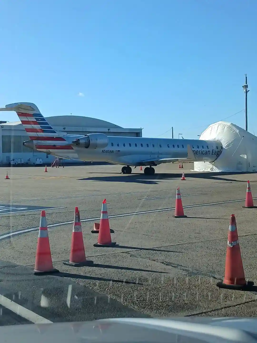 An American Eagle regional jet is parked on an airport tarmac, partially covered by a large white tarp, with orange traffic…