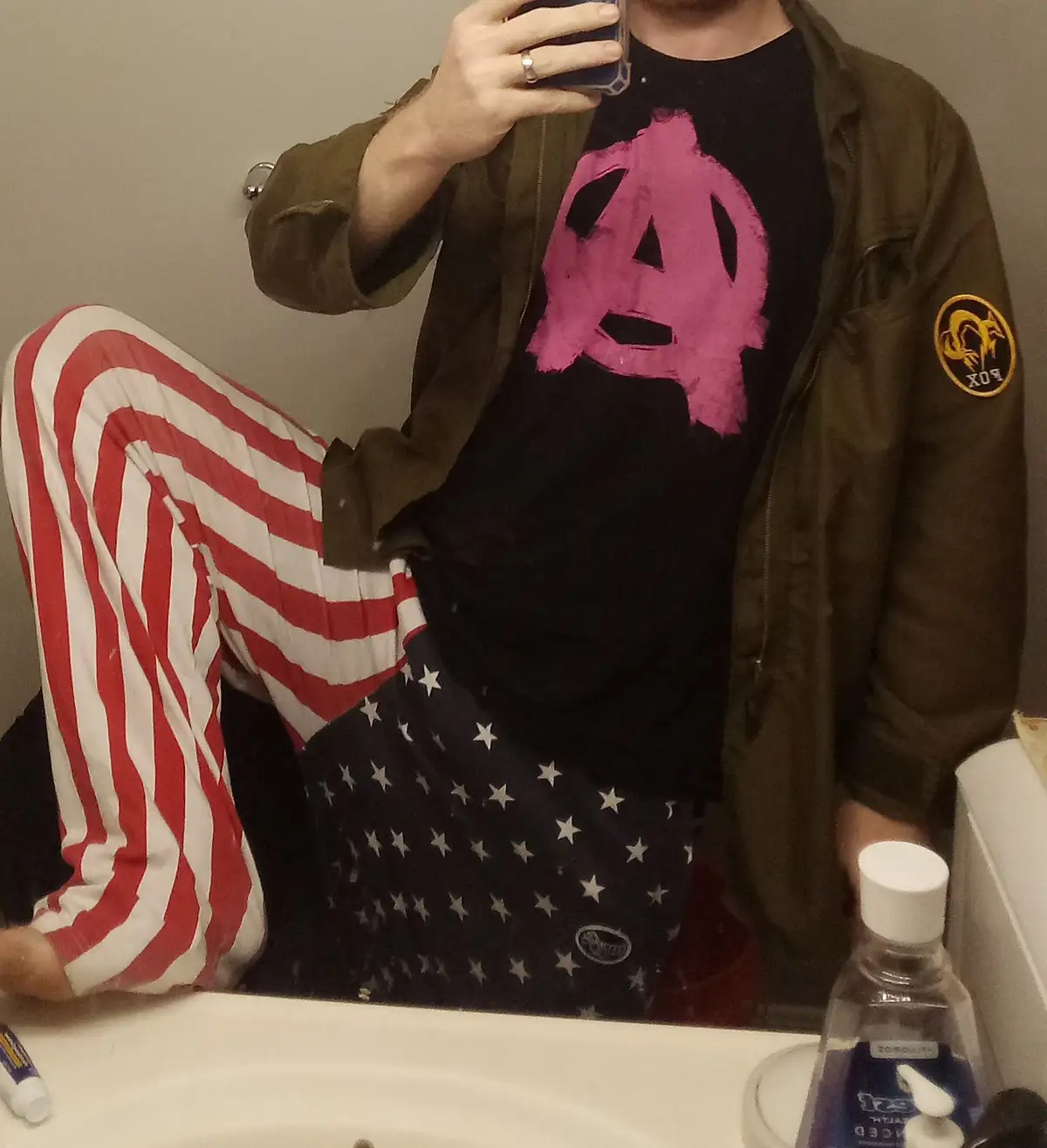 A person takes a mirror selfie in a bathroom, wearing American flag-themed pajama pants, a black t-shirt with a pink…