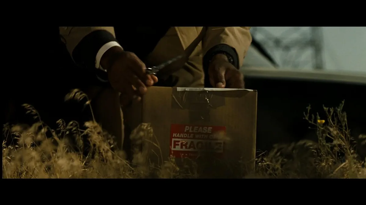 A person in a tan coat uses scissors to open a cardboard box labeled 'PLEASE HANDLE WITH CARE FRAGILE' while sitting in tall…