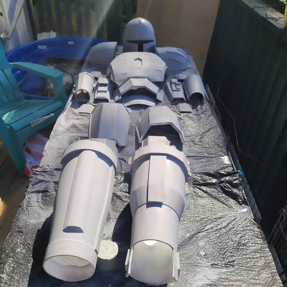 A collection of unpainted, gray 3D-printed armor pieces for a Mandalorian-style costume are laid out on a table covered with…