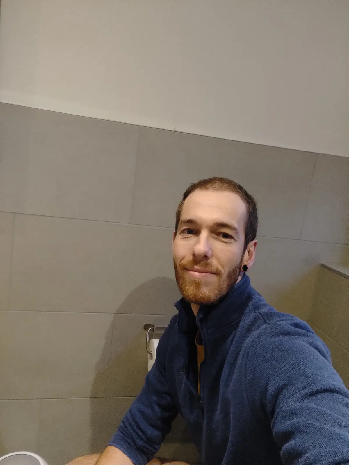 A man with a beard and black ear gauge sits on a toilet in a tiled bathroom, smiling at the camera while holding the phone…