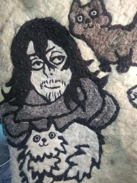 A close-up of a needle-felted artwork depicting a person with long dark hair and a scarf, holding a fluffy white cat, with…