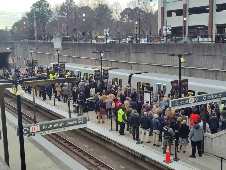 A large crowd of people in business attire stands on a MARTA train platform, lined up to board a modern silver train adorned…
