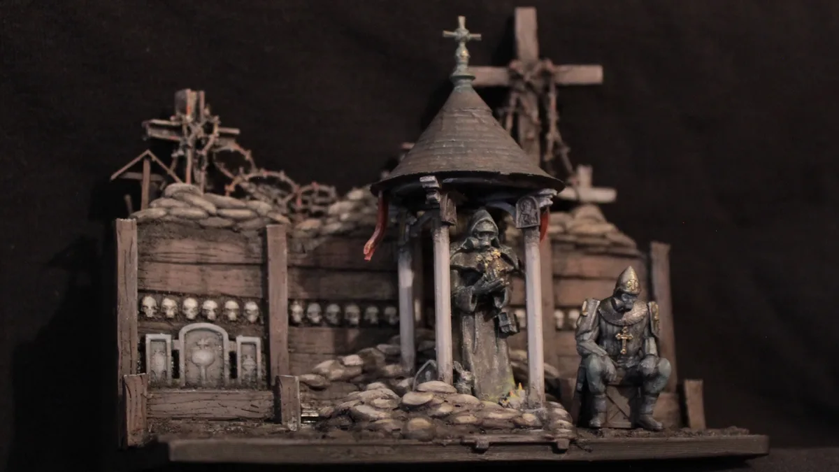 A miniature diorama depicts a grim, war-torn scene with a small chapel, crosses, skulls, and two figures: a robed priest…