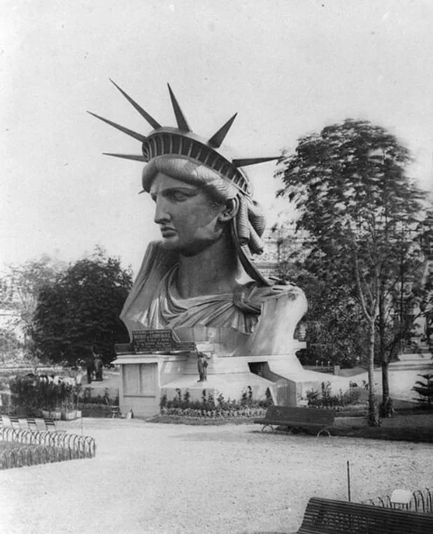 A black-and-white historical photograph shows the head and shoulders of a large statue resembling the Statue of Liberty, set…