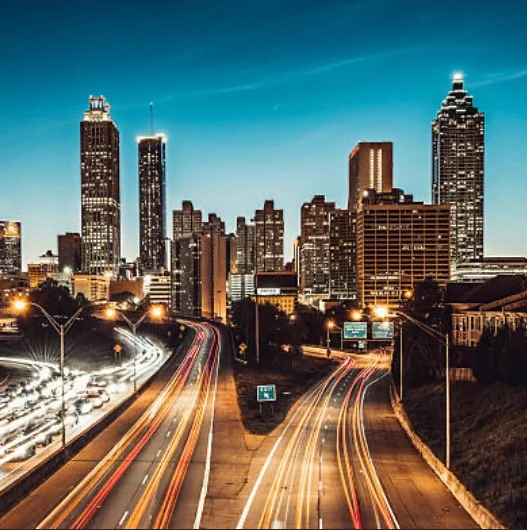 A long-exposure photograph of a city skyline at dusk, showing illuminated skyscrapers and streaks of light from traffic on…
