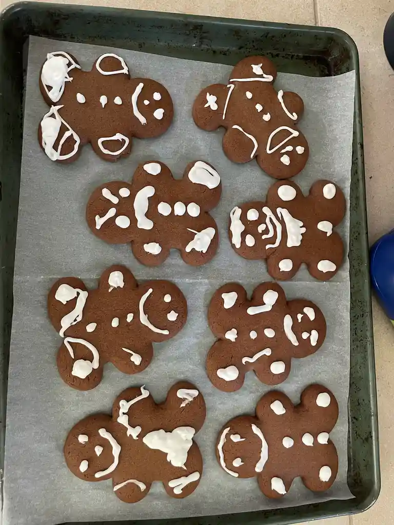 A baking tray lined with parchment paper holds eight gingerbread man cookies, each decorated with white icing in a messy…