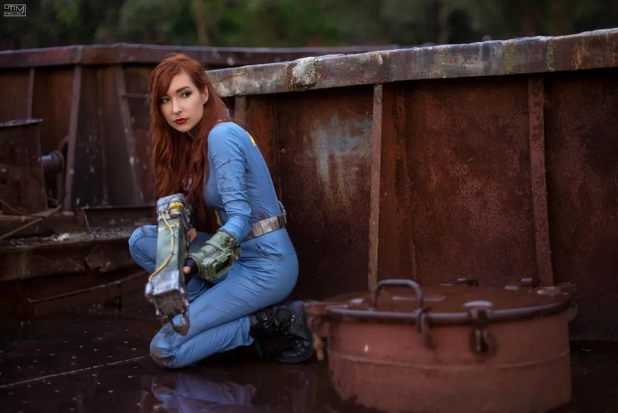 A woman with long red hair, dressed in a blue jumpsuit with a yellow patch and utility belt, kneels beside a rusty metal…