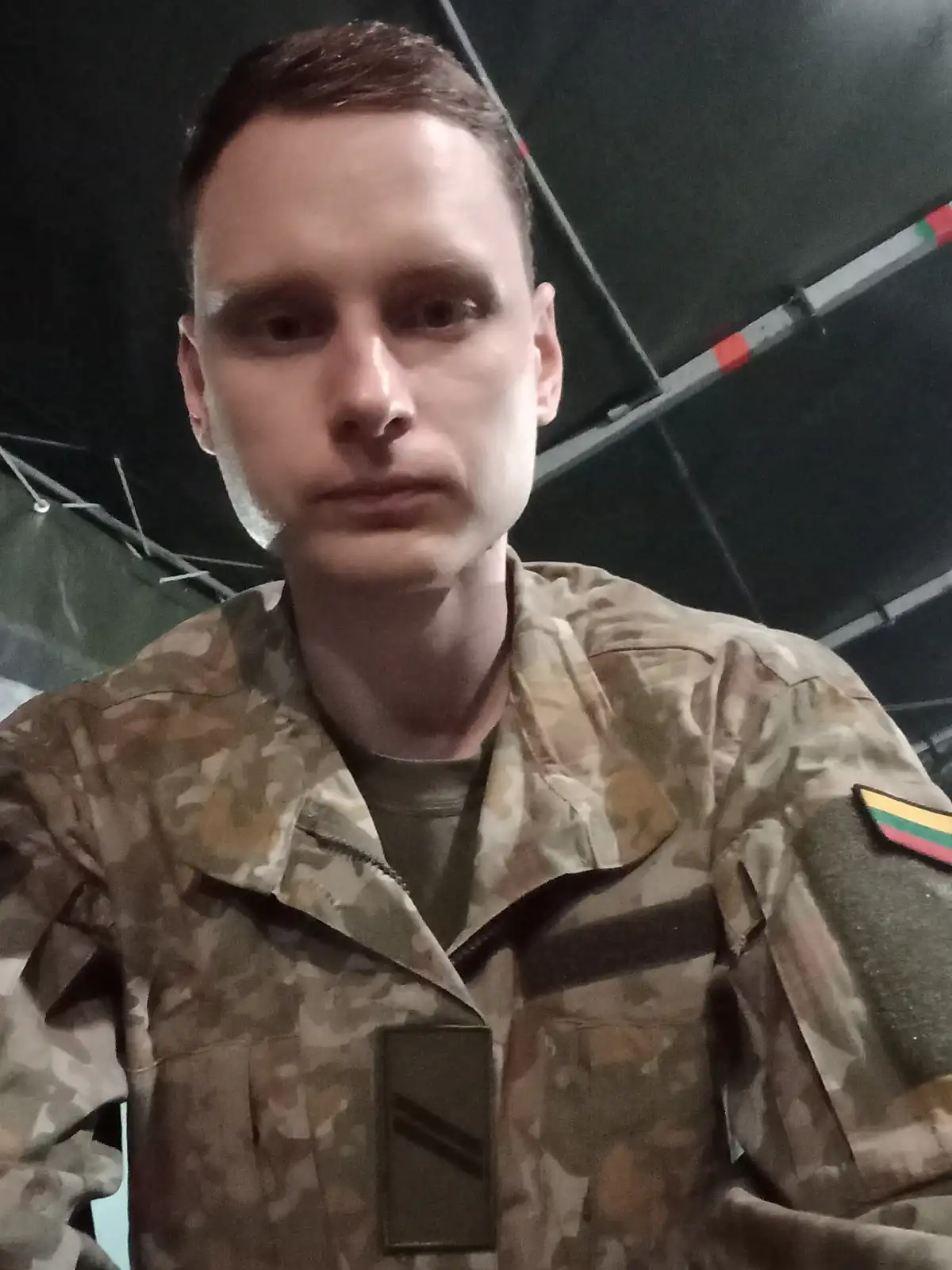 A man in a military camouflage uniform with a Lithuanian flag patch on his right sleeve and a rank insignia on his chest…