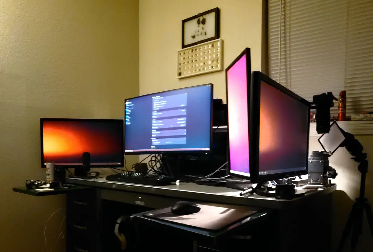 A dimly lit home office desk features four computer monitors arranged in a row, displaying various colorful wallpapers and…