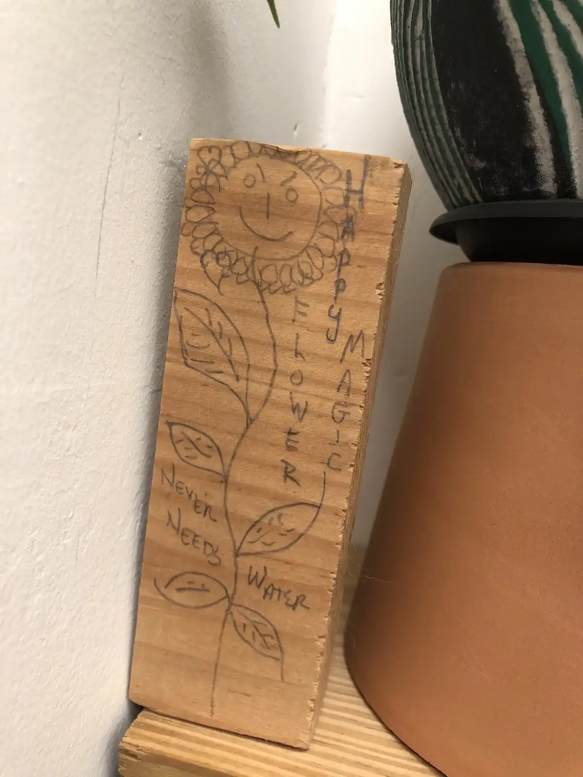 A hand-drawn illustration on a wooden block depicts a smiling sunflower with leaves showing various facial expressions…