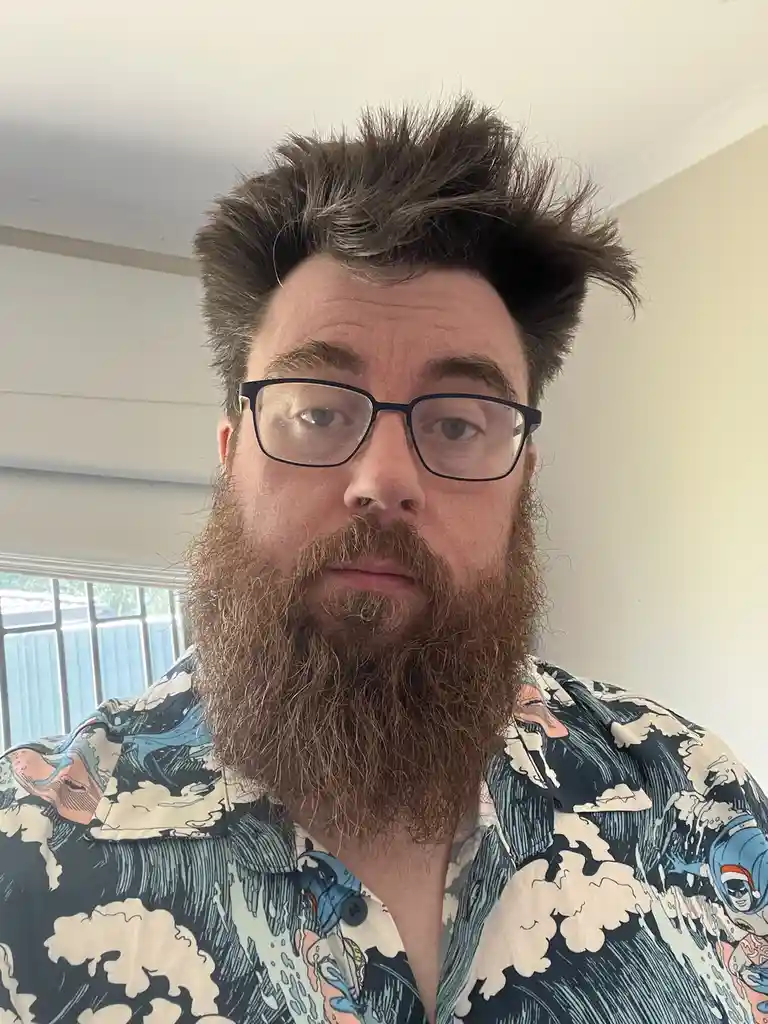 A man with a full brown beard, glasses, and messy brown hair looks directly at the camera with a neutral expression. He…