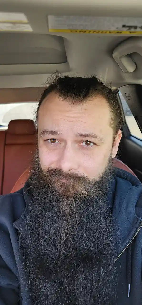 A man with a long, dark beard and brown eyes sits in the driver's seat of a car, looking directly at the camera with a…
