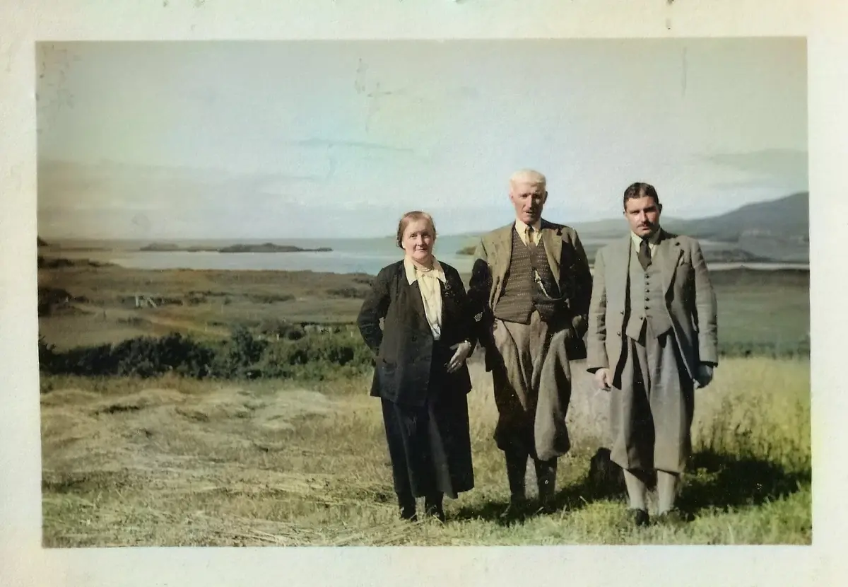 An old, faded color photograph shows three people standing in a grassy field with a body of water and distant hills behind…