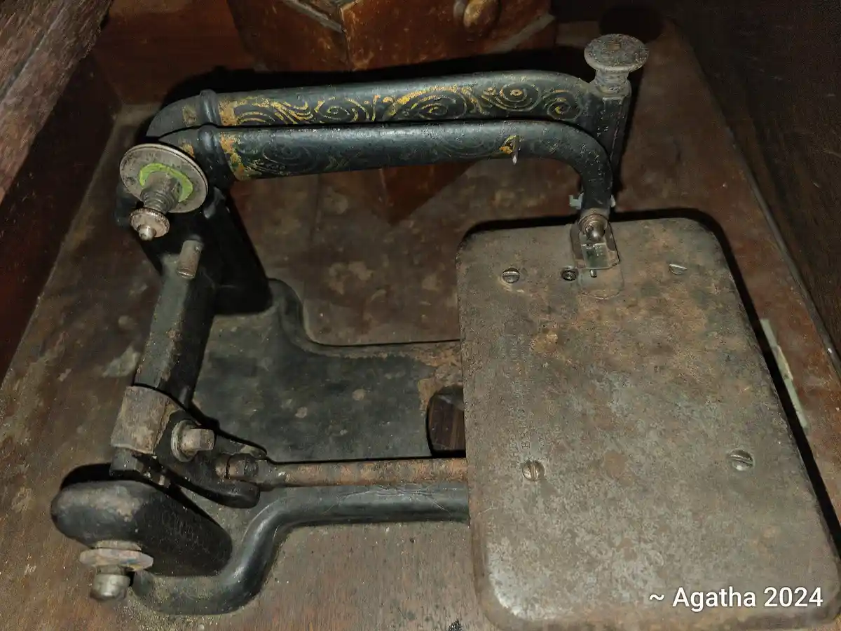 A close-up of a rusty, vintage black treadle sewing machine with faded gold swirl patterns on its arm, resting on a wooden…