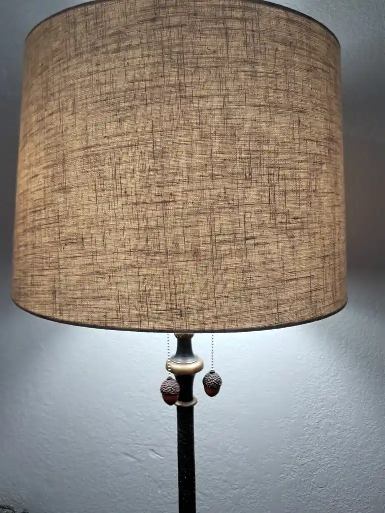 A close-up photo shows a lamp with a beige, textured fabric shade and a dark metal base. Two acorn-shaped pull chains hang…