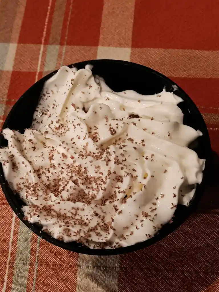 A top-down view of a black bowl filled with white whipped cream, sprinkled with chocolate shavings, resting on a red, beige…