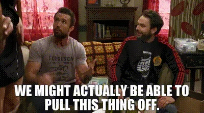 Two men sit on a couch in a living room, reacting to something off-screen. The man on the left wears a grey t-shirt with…