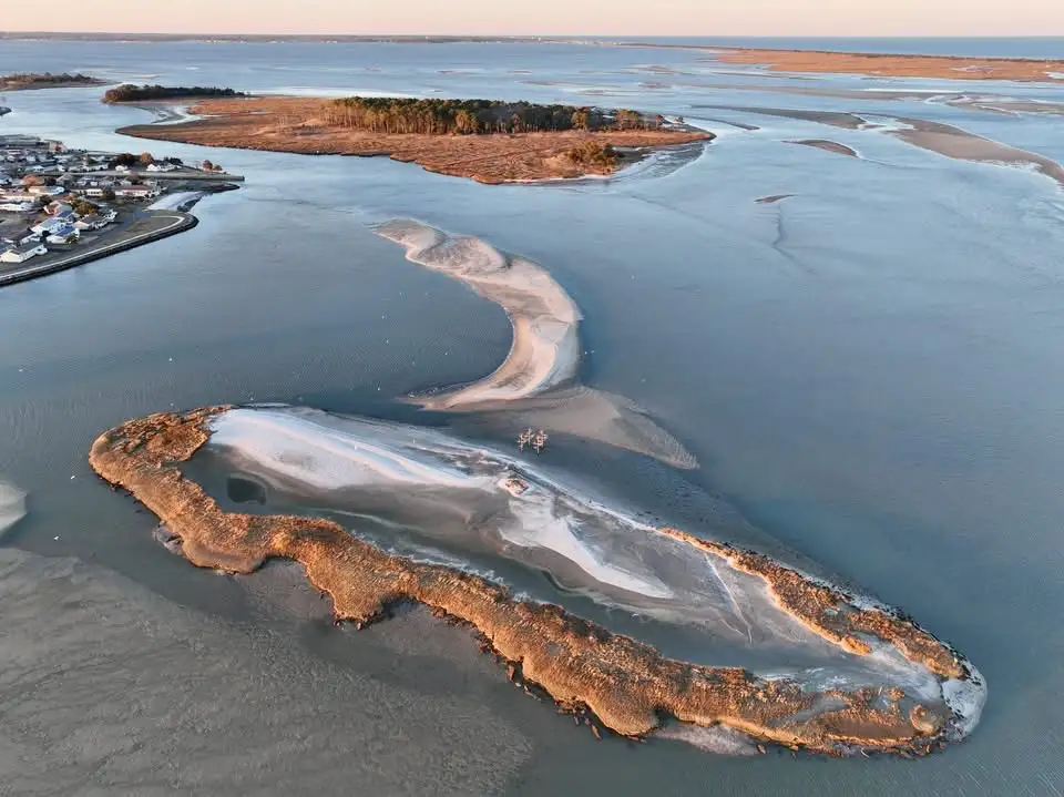 An aerial photograph shows a winding, narrow sandbar or spit of land extending through shallow water, connecting two larger…